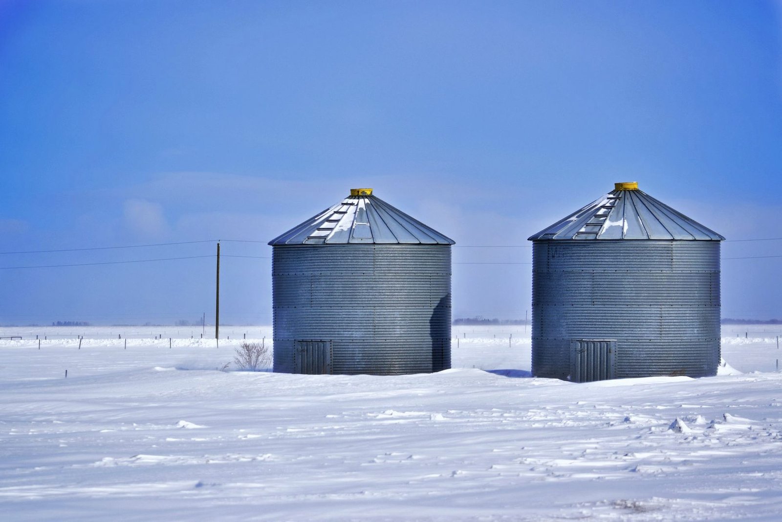 Two corrugated steel grain bins on a snow-covered field under a deep blue sky