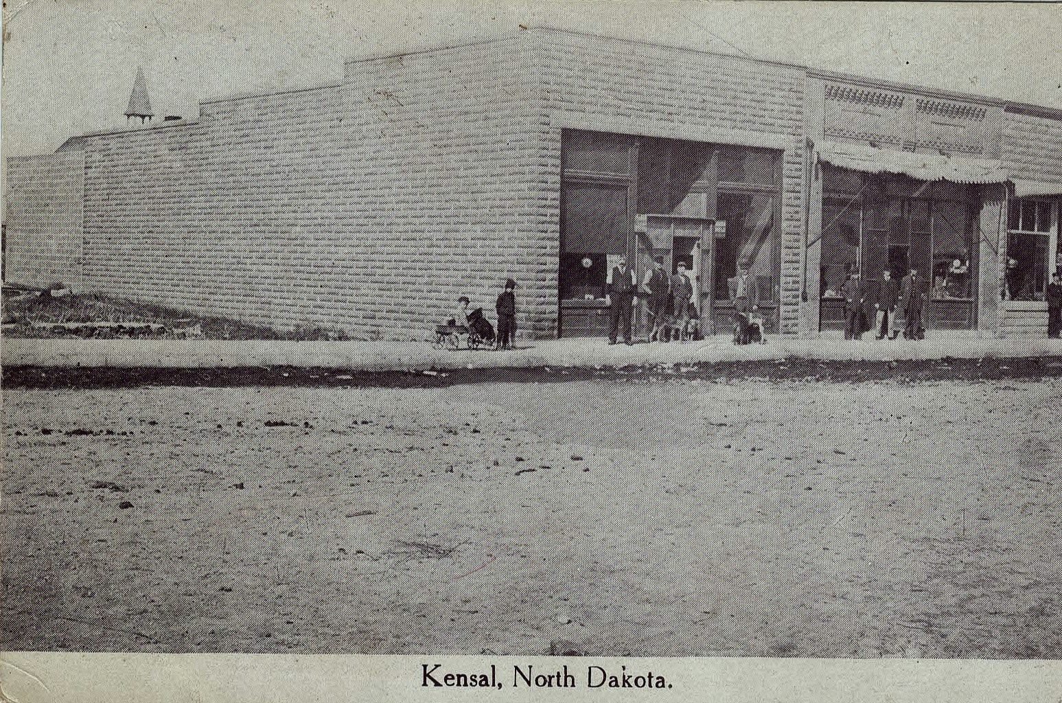 An old postcard of a Kensal street with a brick storefront, labelled Kensal, North Dakota